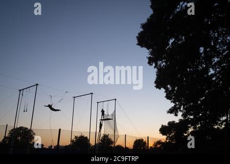 Silhoueté dans le ciel du soir, une figure interprète des acrobaties trapèze à Ruskin Park à Lambeth, le 29 juillet 2020, à Londres, en Angleterre. L'école Trapeze de TLCC arrive à Ruskin Park chaque été, bien qu'au cours de la pandémie du coronavirus, ils aient dû retarder leur présence dans ce sud de London Park. Banque D'Images