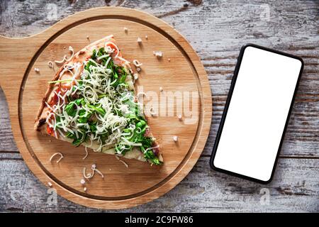 Une délicieuse tranche de pizza avec tomate, salade de roquette et fromage sur un plateau en bois, à côté d'un smartphone avec écran vide et maquette. Capote abaissée Banque D'Images