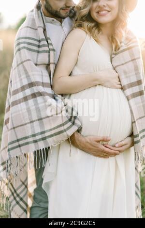 Image rognée d'un jeune couple heureux, d'un homme et d'une femme enceinte, marchant dans une soirée ensoleillée dans un magnifique champ d'été sauvage, recouvert d'une couverture à carreaux Banque D'Images