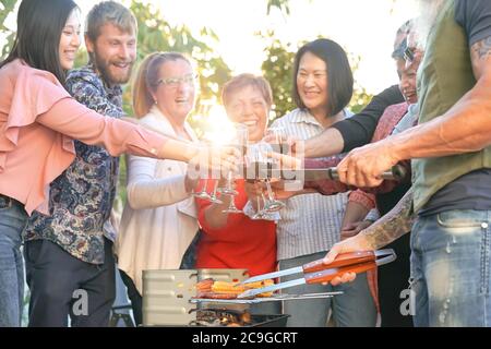 Une famille heureuse qui se réjouit et qui toasque avec du vin rouge lors d'une fête barbecue - chef homme senior qui fait griller de la viande et qui a avec ses parents Banque D'Images