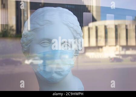 un homme, une femme buste en plâtre dans un masque médical de protection derrière une fenêtre en verre avec un reflet des rues de la ville Banque D'Images