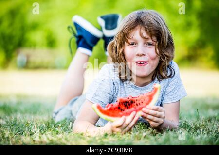 Garçon mangeant du melon d'eau en étant allongé dans le parc, souriant, portrait. Banque D'Images