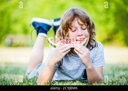 Garçon mangeant du melon d'eau en étant allongé dans le parc, souriant, portrait. Banque D'Images