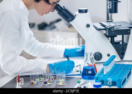Chercheur médical examinant un nouveau médicament. Femme étudiante en sciences vêtue d'une blouse de laboratoire blanche regardant à travers un microscope. Banque D'Images
