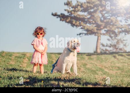 Mignon élégant petite fille caucasienne en lunettes de soleil chien de marche dans le champ de parc à l'extérieur l'été ensoleillé jour. Enfant jouant avec un animal domestique. Banque D'Images