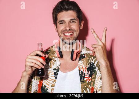 Portrait d'un homme joyeux avec un casque signe de paix tout en buvant du soda isolé sur fond rose Banque D'Images