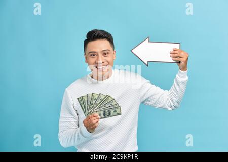 Joyeux jeune homme d'affaires tenant la flèche lots de dollars billets argent liquide isolés sur fond bleu Banque D'Images