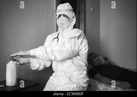 Image en noir et blanc du médecin en combinaison avec l'EPI qui rend visite au patient à la maison, décontaminant les mains avec de l'assainisseur d'alcool avant l'examen du patient. Infirmière portant un masque facial, des gants, regardant la caméra Banque D'Images