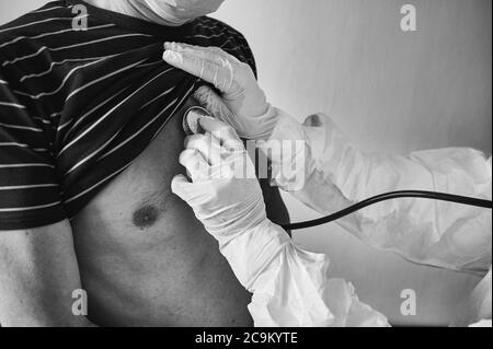 Prise de vue rapprochée d'un médecin en combinaison de protection PPE examinant un patient âgé de sexe masculin à la maison à l'aide d'un phonendoscope, en vérifiant la poitrine, en noir et blanc. Concept de coronavirus Banque D'Images