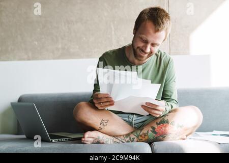 Photo d'un homme souriant à tête rouge avec une barbe qui travaille avec un ordinateur portable et des papiers tout en étant assis sur un canapé à la maison Banque D'Images