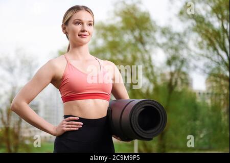 Femme de fitness portant des vêtements de sport tenant un tapis de yoga à l'extérieur dans le parc d'été Banque D'Images