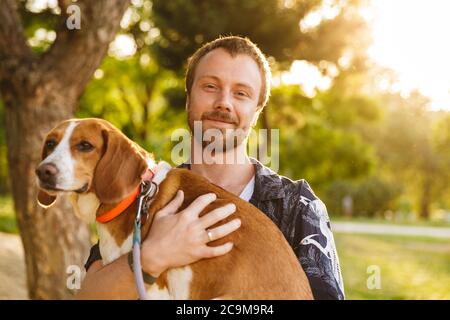 Image d'un jeune homme barbu caucasien souriant tout en tenant son chien beagle entre les mains au parc d'été Banque D'Images