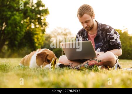 Image d'un jeune homme caucasien barbu utilisant un ordinateur portable tout en étant assis avec son chien beagle sur l'herbe dans le parc d'été Banque D'Images