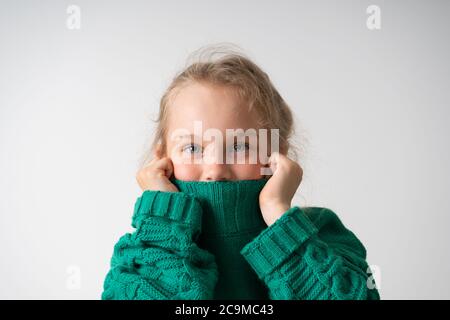 Adorable petite fille cachant la partie inférieure de son visage sous un col épais de pull chaud tricoté. Gros plan studio isolé sur espace de copie blanc Banque D'Images