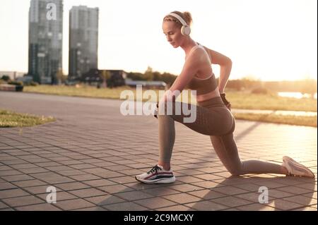 Jeune femme sportive dans le casque qui s'étire dans le parc dans la vue de la ville, jambes fortes et abs, bras musclés, fitness porter fille Banque D'Images