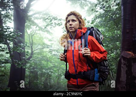 Portrait d'une trekker femelle, portant des vêtements d'extérieur et un sac à dos de randonnée, trekking dans une forêt profonde et brumeuse. Banque D'Images