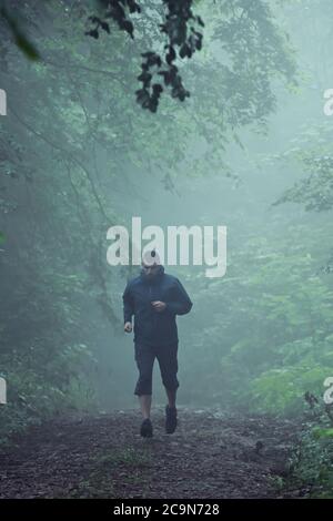 Coureur d'âge moyen, courant à travers le sentier de forêt brumeux, tôt le matin. Banque D'Images