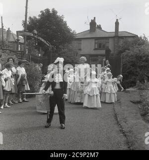 Années 1950, historique, sur un chemin dans un parc de terrain de sport à Farnworth, dans le Lancashire, une adolescente, la « Reine de la Rose » récemment couronnée de la ville, marche avec ses maidens. Les jeunes enfants sont en costumes et un garçon a été deviné comme un crieur de ville. Dans une robe et portant un tiara, la « Reine de la Rose » serait à la tête de la traditionnelle procession ou parade connue sous le nom de « Journée de marche », des célébrations qui étaient communes dans le nord-ouest de l'Angleterre, au Royaume-Uni dans cette époque. Un événement annuel, certains remontent aux années 1830 où ils étaient des défilés d'église liés aux écoles du dimanche des enfants et beaucoup sont encore aujourd'hui. Banque D'Images