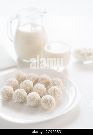 Truffes blanches de noix de coco sucrées sur une assiette, verre de lait et carafe, fromage cottage dans un bol sur fond défoqué, sur une table blanche, dans une clé haute. Banque D'Images