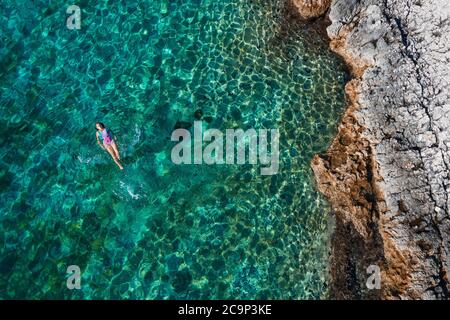 Vue aérienne de haut de la jeune femme flottant à l'arrière et de détente sur les chaudes eaux turquoise de la mer Adriatique avec la côte rocheuse. Des gens insouciants v