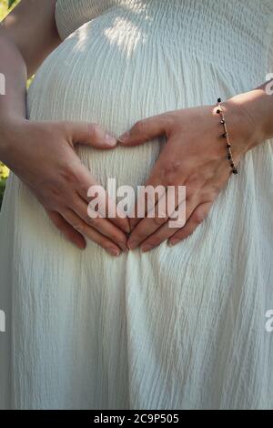 Gros plan mignon composition avec une jeune femme enceinte portant une robe blanche fait un symbole en forme de coeur avec ses mains sur son ventre. Bonne mère attendant Banque D'Images