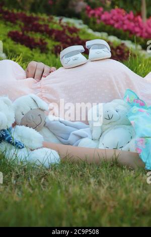 Composition mignonne avec une jeune femme enceinte qui s'est posée à l'extérieur sur l'herbe dans le parc, entourée de nombreux jouets en peluche et de chaussures pour nouveau-né Banque D'Images