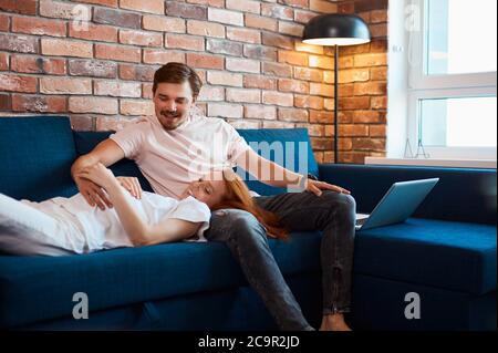 couple caucasien marié, s'attendant à bébé, se reposent sur un canapé ensemble. le jeune homme aime sa femme à tête rouge, à la maison en tenue domestique Banque D'Images