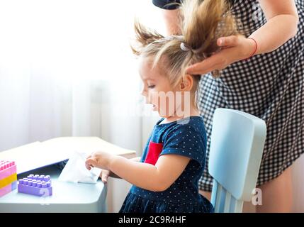 Crop femme faisant des queues de cheval pour adorable petite fille assise à la table et jouant avec des jouets pendant le week-end à la maison Banque D'Images