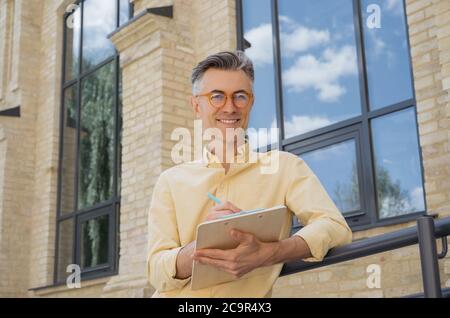 Projet de travail d'homme d'affaires senior, planification de démarrage, regarder la caméra. Portrait d'un beau écrivain portant des lunettes élégantes prenant des notes Banque D'Images