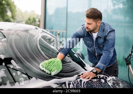 Beau jeune homme caucasien nettoyant son pare-brise de voiture avec une mitaine verte et de la mousse de savon dehors au service de lavage de voiture Banque D'Images