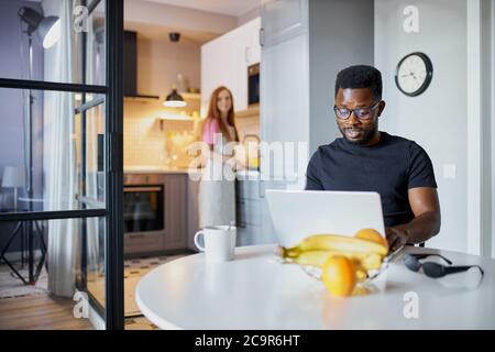 couple interracial dans la cuisine, le matin. femme redhead parle avec le mari noir travaillant pendant la cuisine Banque D'Images