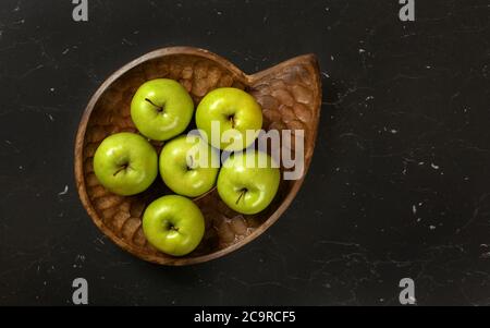 Vue de dessus en bas, six pommes vertes dans un bol en bois sculpté, sur un plateau en marbre noir. Banque D'Images