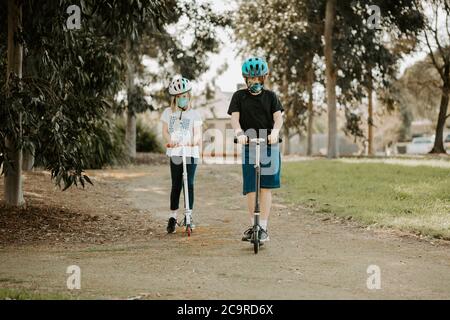 Deux enfants caucasions qui portent des scooters et portent des masques pendant la pandémie Corona COVID-19 Banque D'Images