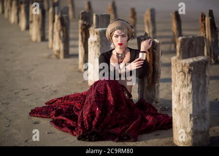 la jeune fille de la danseuse est assise sur la terre et penche les coudes sur les colonnes en bois entraînées dans le sol et regarde dans un coup de feu en robe longue rouge avec un make-u. Banque D'Images