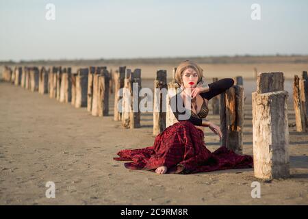 la jeune fille de la danseuse est assise sur la terre et penche les coudes sur les colonnes en bois entraînées dans le sol et regarde dans un coup de feu en robe longue rouge avec un make-u. Banque D'Images