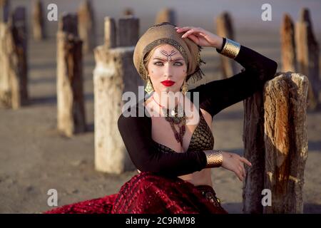 le portrait de fille de la danseuse est assis sur la terre et penche les coudes sur les colonnes en bois conduit dans le sol et regardez dans une photo dans la robe longue rouge avec Banque D'Images