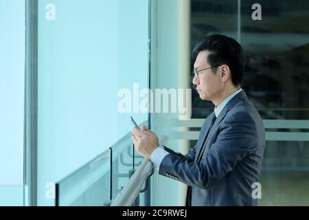 Homme d'affaires chinois en costume debout avec les mains tenant son téléphone portable. Vue latérale. Banque D'Images