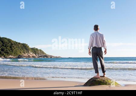 Homme d'affaires mûr appréciant la plage tout en se tenant seul sur un rocher au lever du soleil Banque D'Images