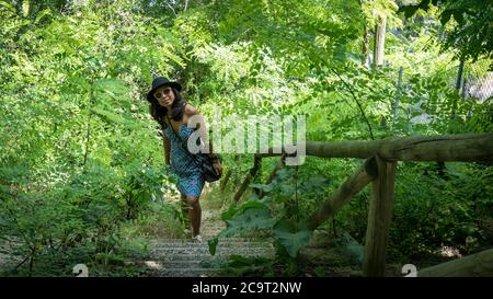 Femme asiatique avec lunettes de soleil, robe bleue et chapeau noir debout au bas des escaliers avec main courante en bois brut à Bologne Italie Banque D'Images