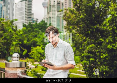 Homme d'affaires ou étudiant dans une robe décontractée utilisant une tablette un parc tropical sur le fond de gratte-ciel. Robe de chambre blanche, short beige Banque D'Images