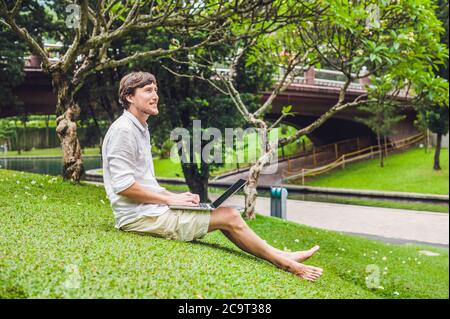 Homme d'affaires ou étudiant en robe décontractée utilisant un ordinateur portable dans un parc tropical sur le fond de gratte-ciel. Robe de chambre blanche, short beige Banque D'Images