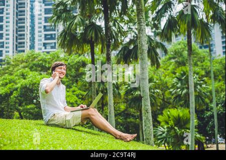 Homme d'affaires ou étudiant en robe décontractée utilisant un ordinateur portable dans un parc tropical sur le fond de gratte-ciel. Robe de chambre blanche, short beige Banque D'Images