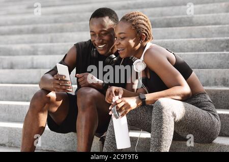 Après la formation. Heureux couple africain se reposant sur les escaliers à l'extérieur et en utilisant un smartphone Banque D'Images