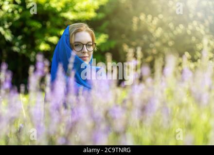 Portrait d'une jeune femme souriante avec des lunettes et une tête portant un foulard bleu pose derrière la lavande dans le jardin Banque D'Images