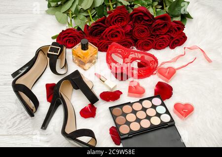 Masque de carnaval rouge, parfum, chaussures et rouge à lèvres sur une table en bois sur un fond de roses de scarlet Banque D'Images