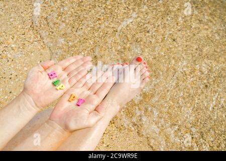 Les mains et les pieds des femmes avec une pédicure rouge sur fond de sable de mer et de surf. Dans les paumes des lettres avec l'inscription Happy Banque D'Images