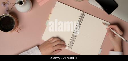 Photo de la femme qui écrit sur un carnet vierge sur une table rose avec fournitures et décorations Banque D'Images