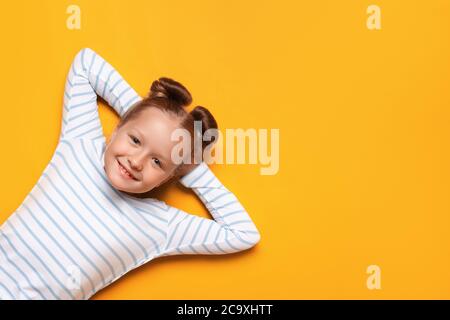 Portrait d'une jolie petite fille adorable et adorable avec des petits pains à cheveux sur fond jaune. L'enfant est allongé avec ses mains repliées derrière sa tête. Fermer- Banque D'Images