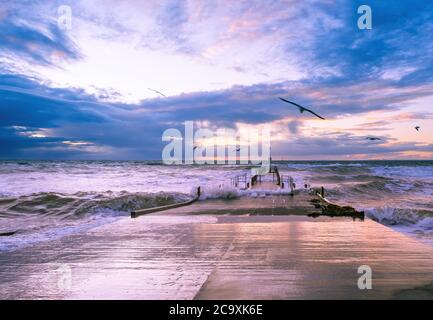 Des mouettes survolant de grandes vagues écrasant sur une petite jetée au coucher du soleil Banque D'Images