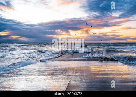 Vagues de tempête écrasant sur un quai avec des mouettes volantes au coucher du soleil à Melbourne, en Australie Banque D'Images